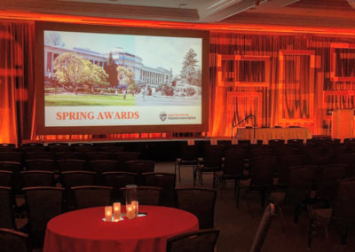 Chairs are arranged in front of a stage with a large screen reading "Spring Awards" at a OneSource Strategy alumni event