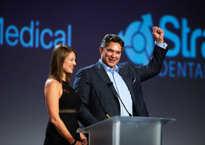 A Man and Woman Speaking at a Podium on Stage at a OneSource Strategy Event, The Man Raising His Fist in the Air