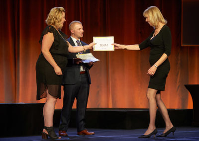 A Woman Receiving a Certificate on Stage at a OneSource Strategy Event, While a Man is Speaking