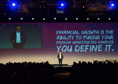 A Man Speaking Onstage at a OneSource Strategy Event With Text on a Large Screen Behind Him Reading "Financial Growth is the Ability to Pursue Your Passion Unrestricted However You Define It."