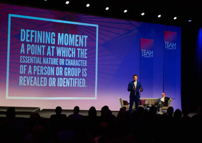 A Man Speaking Onstage at a OneSource Strategy Event With Text on a Large Screen Behind Him Reading "Defining Moment: A Point at Which the Essential Nature or Character of a Person or Group is Revealed or Identified.""