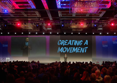 A Wide Shot of a Man Speaking Onstage at a OneSource Strategy Event With Text on a Large Screen Behind Him Reading "Creating a Movement"