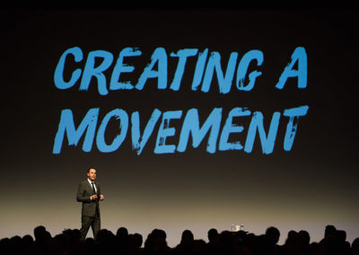 A Man Speaking Onstage at a OneSource Strategy Event With Text on a Large Screen Behind Him Reading "Creating a Movement"