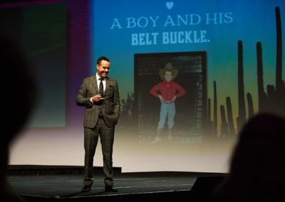 A Man Speaking Onstage at a OneSource Strategy Event in Front of a Large Screen Behind Him With Text Reading "A Boy and His Belt Buckle." Above a Photo of a Child in a Cowboy Costume