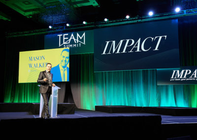 A Man Speaking Onstage at a OneSource Strategy Event in Front of a Large Screen With Text Reading "IMPACT"