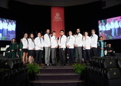 A line of graduates in white coats on stage at a OneSource Strategy higher education event
