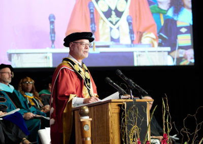 A man in graduation cap and gown speaks at a podium at a OneSource Strategy higher education ceremony