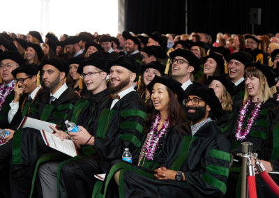 A crowd of graduates caps and gowns laugh at a speech during a OneSource Strategy higher education ceremony