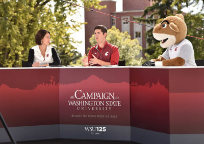 A man, woman, and cougar mascot speak at an outdoors presentation panel at a OneSource Strategy higher education event