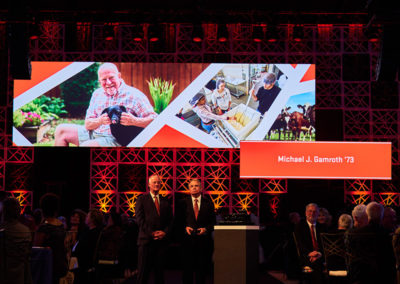 Two men in suits stand next to a collection of gifts for honorees at a OneSource Strategy events