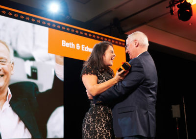 A woman excitedly presents a small black box with an orange ribbon to a man onstage at a OneSource Strategy higher education honoree event