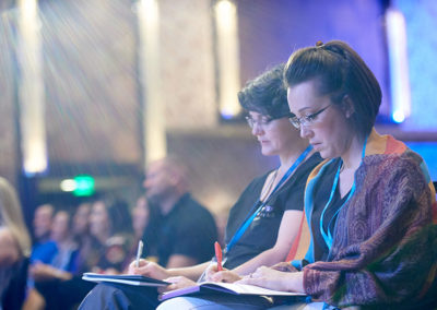 Two people in an audience take notes during training at a OneSource Strategy association event